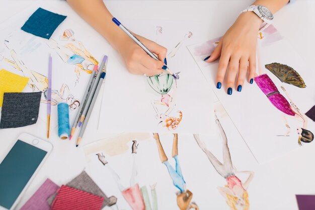 This picture describes the processes of designing clothes. There are hands of a girl drawing sketches on the table. There is creative mess with different stuff on the table.