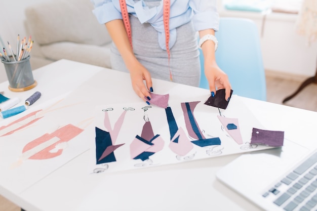 Questa immagine descrive i processi di progettazione di abiti in officina. ci sono mani di una ragazza che cercano gli schizzi e i materiali sul tavolo.