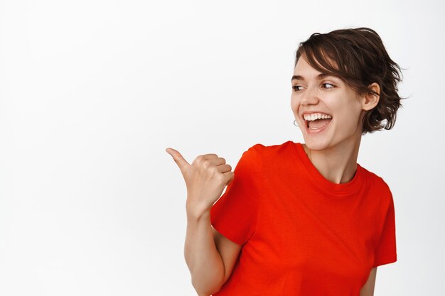 This way. Beautiful modern girl laughing, pointing thumb left and looking at copy space with happy smile on white