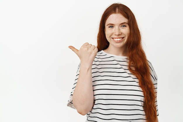 This way. Young happy redhead woman with freckles, ginger girl pointing thumb left and smiling on white