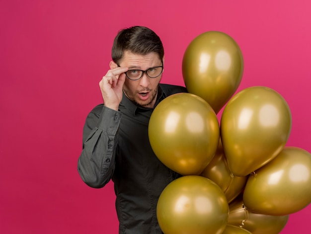 Free photo thoughful young party guy wearing black shirt holding balloons wearing and grabbed glasses isolated on pink