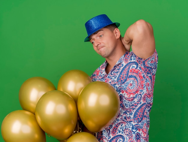 Free photo thoughful young party guy wearing blue hat holding and looking at balloons and putting hand behind head isolated on green