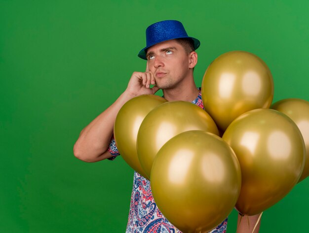 thoughful young party guy wearing blue hat standing among balloons isolated on green