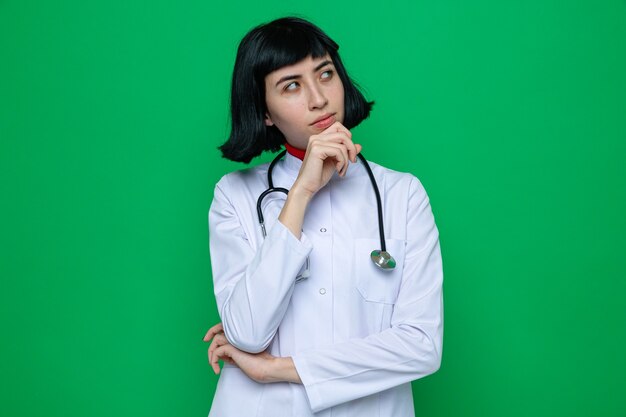 Thoughful young pretty caucasian girl in doctor uniform with stethoscope putting hand on chin and looking at side 