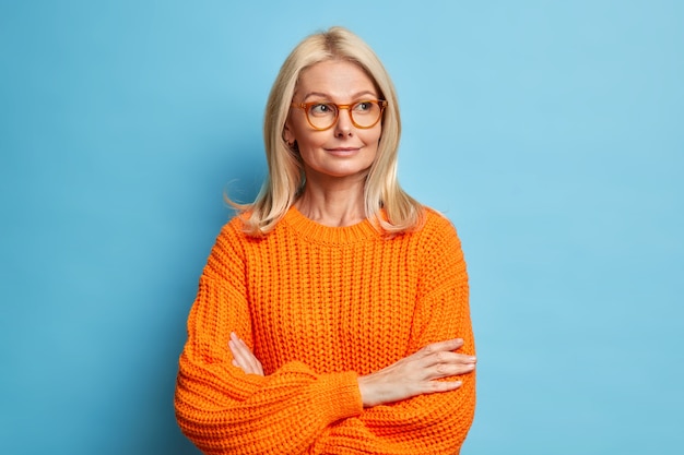 思いやりのある愛らしい金髪の40歳の喜んでいる女性は腕を組んで何かを考えて目をそらし、眼鏡ニットのセーターを着ています。