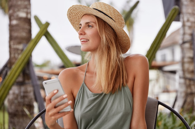 Thoughtful adorable woman in casual clothing, keeps modern smart phone, looks aside, recreate outdoor during summer weather, dreams about something pleasant. People, technology, lifestyle concept