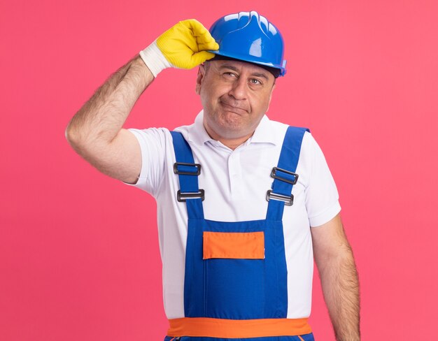 Thoughtful adult builder man in uniform wearing protective gloves holds safety helmet isolated on pink wall