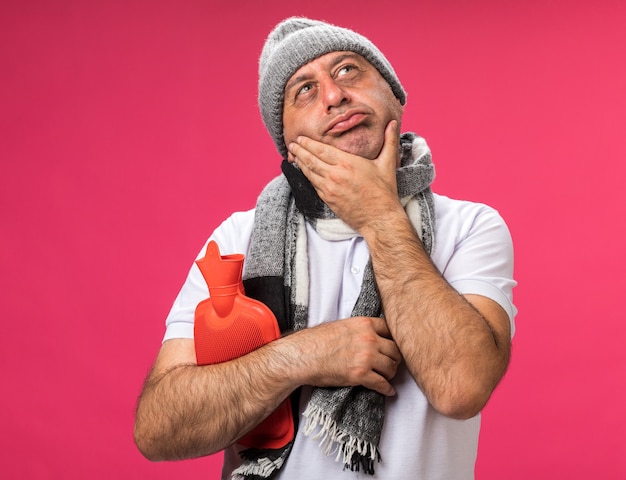 thoughtful adult ill caucasian man with scarf around neck wearing winter hat puts hand on chin and holds hot water bottle looking up isolated on pink wall with copy space