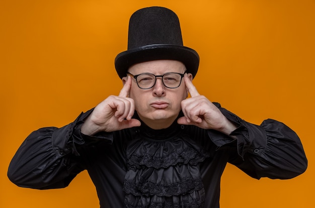 Thoughtful adult man with top hat and glasses in black gothic shirt putting fingers on his temples and looking 