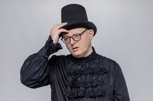 Thoughtful adult man with top hat and glasses in black gothic shirt putting hand on his hat and looking at side 