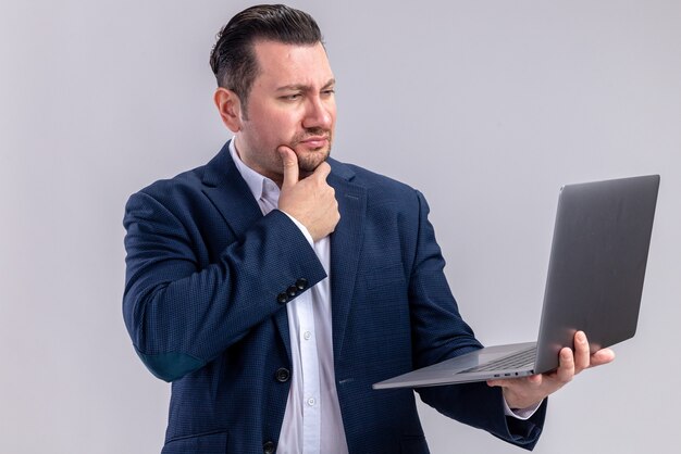 Thoughtful adult slavic businessman holding and looking at laptop isolated on white wall with copy space
