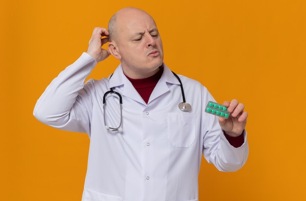 Free photo thoughtful adult slavic man in doctor uniform with stethoscope holding and looking at medicine blister pack