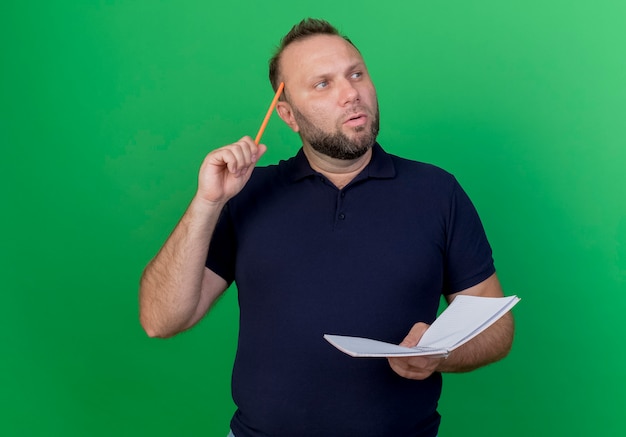 Uomo slavo adulto premuroso che esamina la matita e il rilievo di nota della tenuta del lato toccando la testa con la matita isolata sulla parete verde