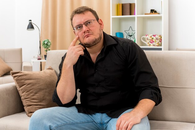Thoughtful adult slavic man in optical glasses sits on armchair putting finger on face looking at side inside the living room
