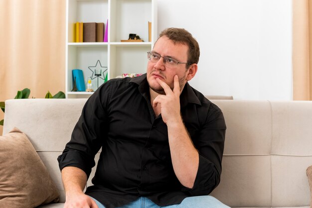 Thoughtful adult slavic man in optical glasses sits on armchair putting hand on chin inside the living room