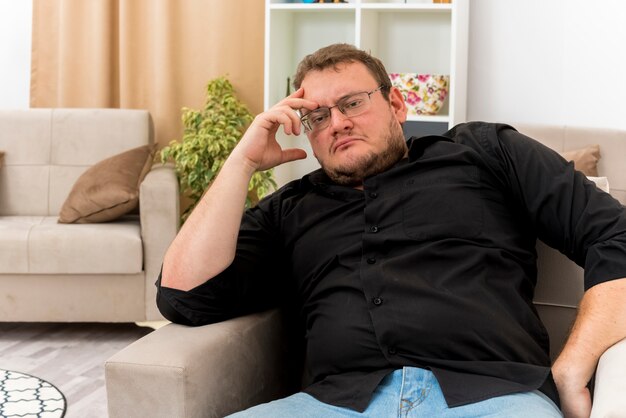 Thoughtful adult slavic man in optical glasses sits on armchair putting hand on head inside living room
