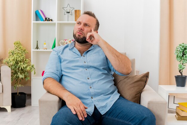 Thoughtful adult slavic man sits on armchair putting finger on temple looking at side inside the living room