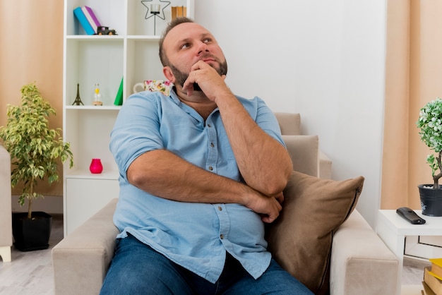 Thoughtful adult slavic man sits on armchair putting hand on chin looking up inside the living room