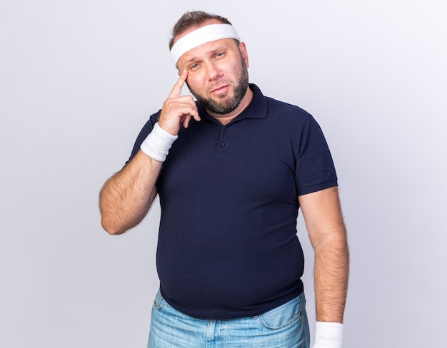 Uomo sportivo slavo adulto premuroso che indossa la fascia e braccialetti che mettono il dito sul tempio isolato sul muro bianco con lo spazio della copia