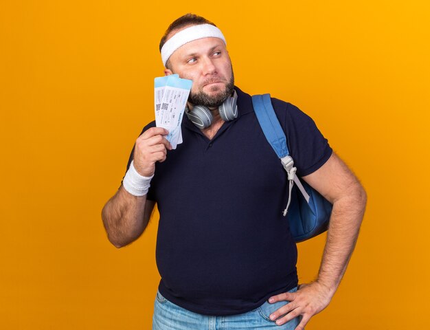 thoughtful adult slavic sporty man with headphones wearing backpack headband and wristbands holding air tickets and looking at side isolated on orange wall with copy space