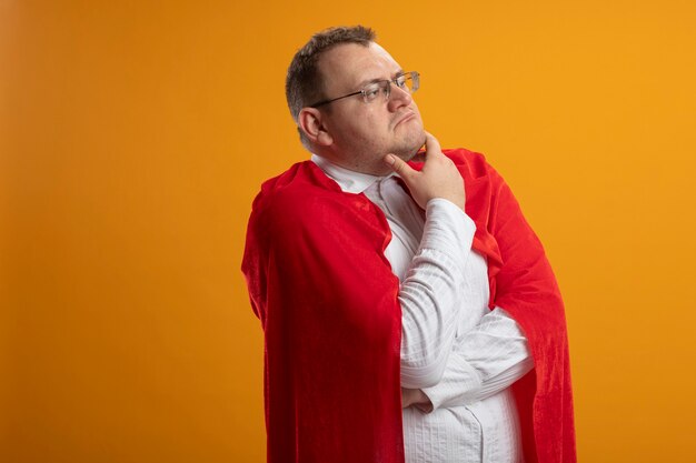 Thoughtful adult slavic superhero man in red cape wearing glasses standing with closed posture looking straight isolated on orange background with copy space