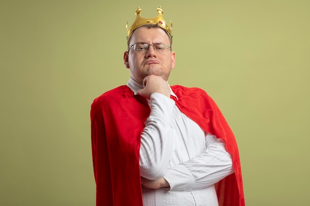 Foto gratuita uomo adulto premuroso del supereroe in mantello rosso con gli occhiali e corona che mette la mano sotto il mento guardando la parte anteriore isolata sulla parete verde oliva