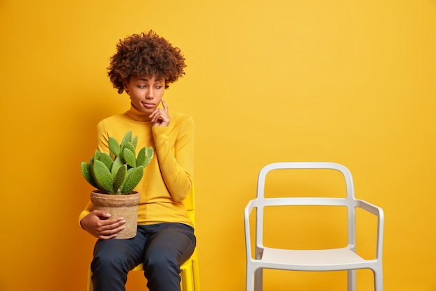 Thoughtful Afro American woman concentrated pensively on empty chair holds potted cactus feels lonely wears casual clothing