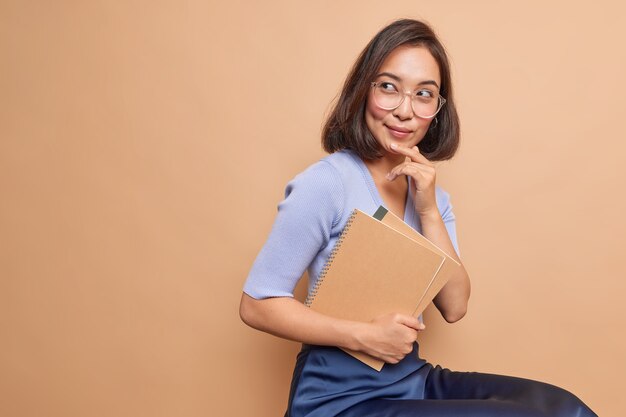 Thoughtful Asian female schoolgirl carries spiral notebooks returns back to school thinks how to improve her knowledge wears spectacles casual clothes sits indoor blank copy space on beige wall