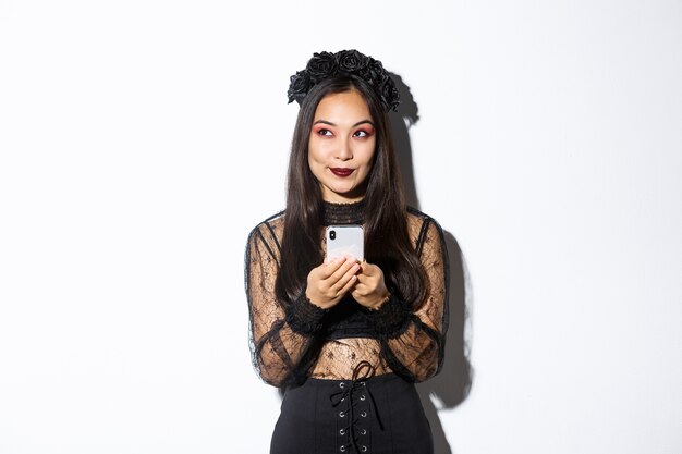 Thoughtful asian girl in elegant dress holding smartphone while thinking, looking at upper left corner, standing over white background.