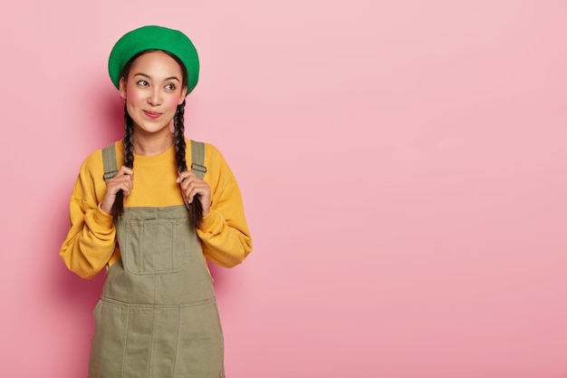 Free photo thoughtful asian lady daydreams about something, touches braids, dressed in stylish outfit, has spare time