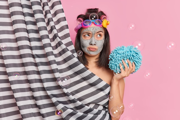 Задумчивая азиатская женщина применяет глиняную маску, делает прическу с роликами для волос, держит губку для душа, имеет глубокие мысли, принимая позы для душа за занавеской, позирует на розовом студийном фоне