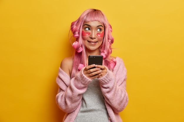 Thoughtful Asian woman gets hair styled, applies curlers, beauty collagen patches, holds mobile phone in hands