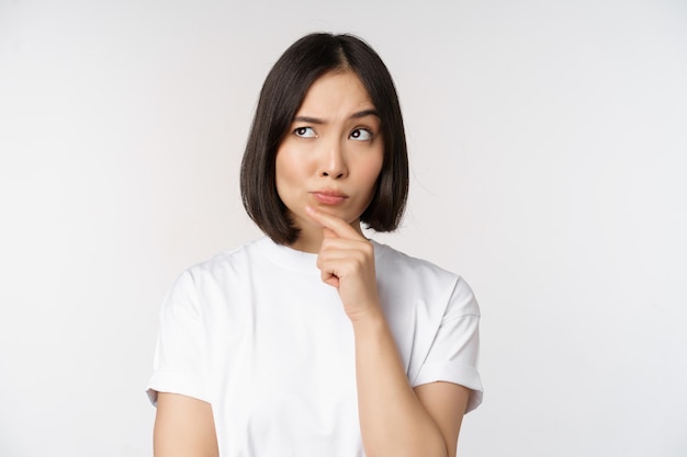 思いやりのあるアジアの女性は、白い背景の上に立っている仮定を考えたり、smthを選択したりすることを熟考して脇を探しています