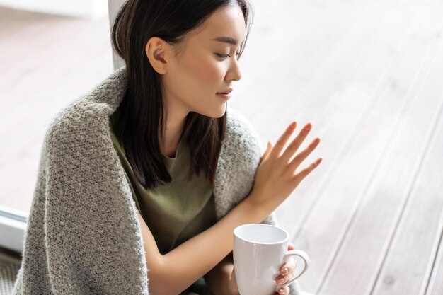 사려 깊은 아시아 여성이 집에 앉아 유리창을 만지며 외출을 갈망합니다.