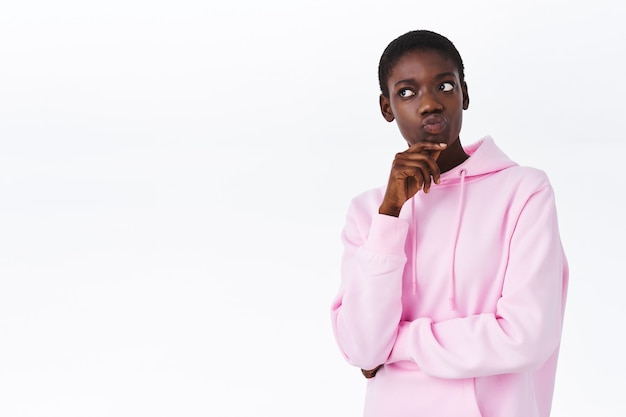 Thoughtful attractive african-american female with short hair, wear pink stylish hoodie, pouting and looking left curious, thinking