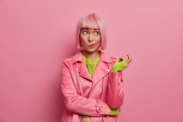 Thoughtful attractive Asian female looks away and thinks hesitant, purses lips and raises hand, tries to decide how to waste money, dyed rosy bob hair