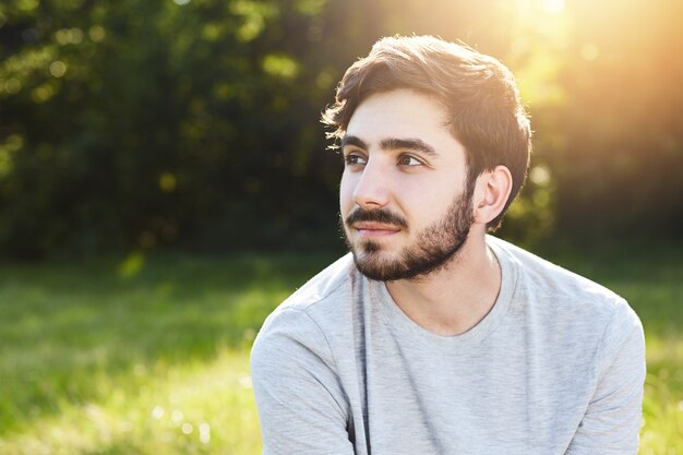 Thoughtful attractive male with dark mustache and beard looking aside into distance dreaming about something pleasant while relaxing