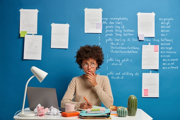 Free photo thoughtful attractive woman in spectacles writes in diary to do list of goals, makes notes in personal organizer