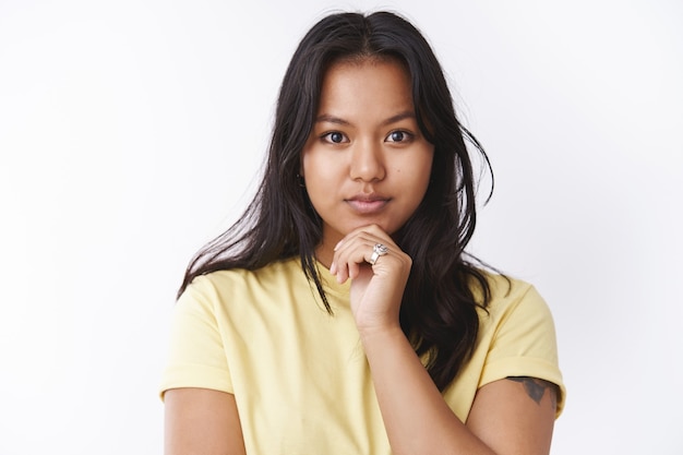 Thoughtful attractive young malaysian girl with facial scars and acne touching chin and looking determined at camera, thinking standing dreamy against white background in yellow t-shirt