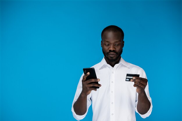 思慮深いひげを生やしたアフロアメリカンの男は携帯電話を保持し、クレジットカードを探しています