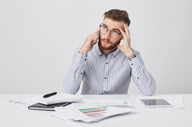 Thoughtful bearded fashionable man works in office, surrounded with graphics and modern devices