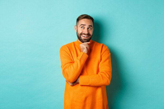 Thoughtful bearded guy making choice, shopping and looking at upper left corner, smiling satisfied, standing over turquoise wall.