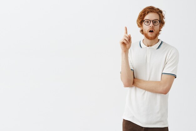 Thoughtful bearded redhead guy posing against the white wall with glasses