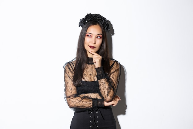 Thoughtful beautiful asian woman standing in black lace dress and gothic wreath, looking upper left corner intrigued. Girl in halloween costume making choice.