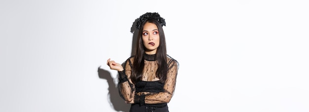 Thoughtful beautiful asian woman in witch costume biting lip and looking upper left corner, daydreaming while standing in halloween widow dress, standing over white background