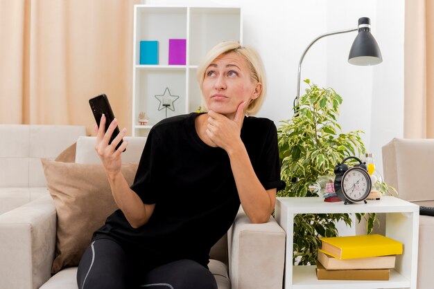 Thoughtful beautiful blonde russian woman sits on armchair putting hand on chin and holding phone looking up inside living room
