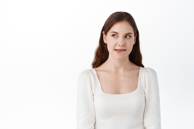 Thoughtful beautiful girl wants taste something tasty biting lip from temptation looking aside at intriguing thing standing pensive against white background