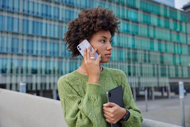 곱슬머리를 한 사려깊은 아름다운 여성이 휴대형 태블릿을 들고 전화 통화를 하며 연락을 유지하기 위해 현대적인 가제트를 사용하여 도심 기술 개념에서 녹색 스웨터 포즈를 취합니다.
