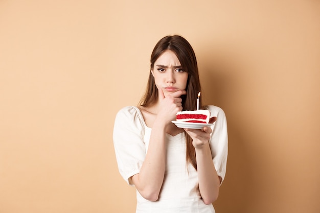 Thoughtful birthday girl frowning and thinking of wish holding cake with candle standing on beige ba...