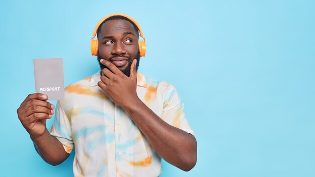 Thoughtful black bearded man holds chin looks away holds passport which contains basic identifying information listens music via wireless headphones isolated over blue wall